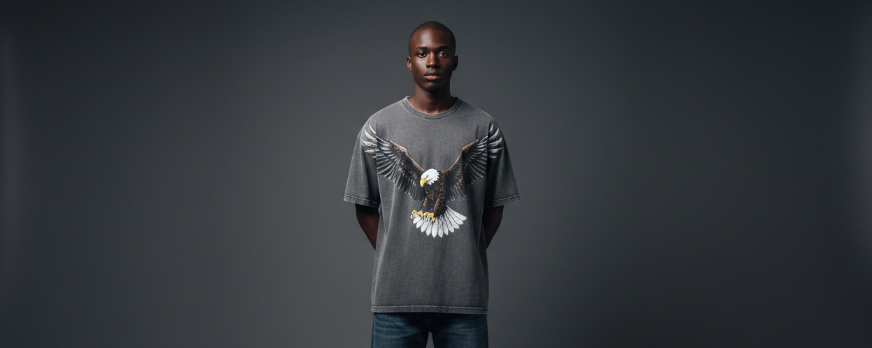 straight faced black male model with no facial hair wearing a washed grey oversized t-shirt with a large eagle image on the front. t-shirt not tucked in. both hands behind his back. full body shot. dark grey background. model standing straight